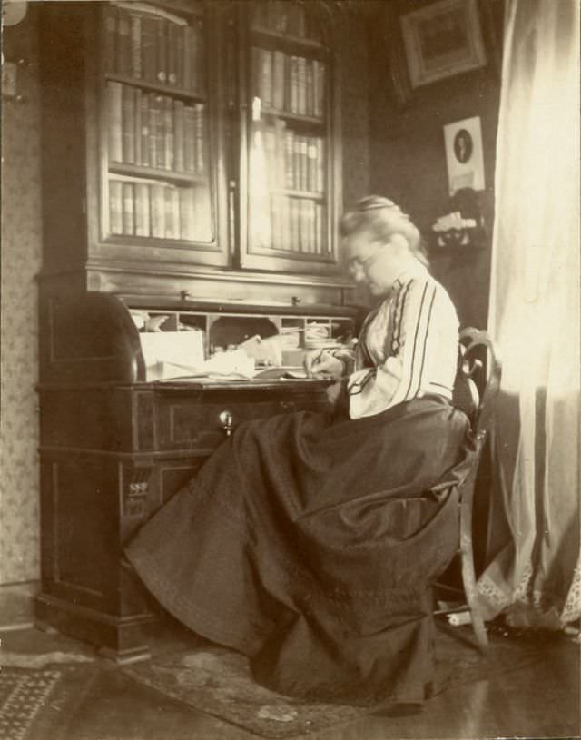 Katherine Horack, pen in hand, seems to be writing at a desk in the sitting room, 1890s