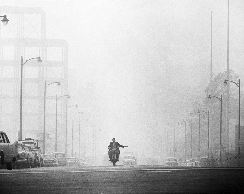 A motorcyclist in Los Angeles prepares to turn while driving along a street which is engulfed in a thick haze combined by fog and smog.