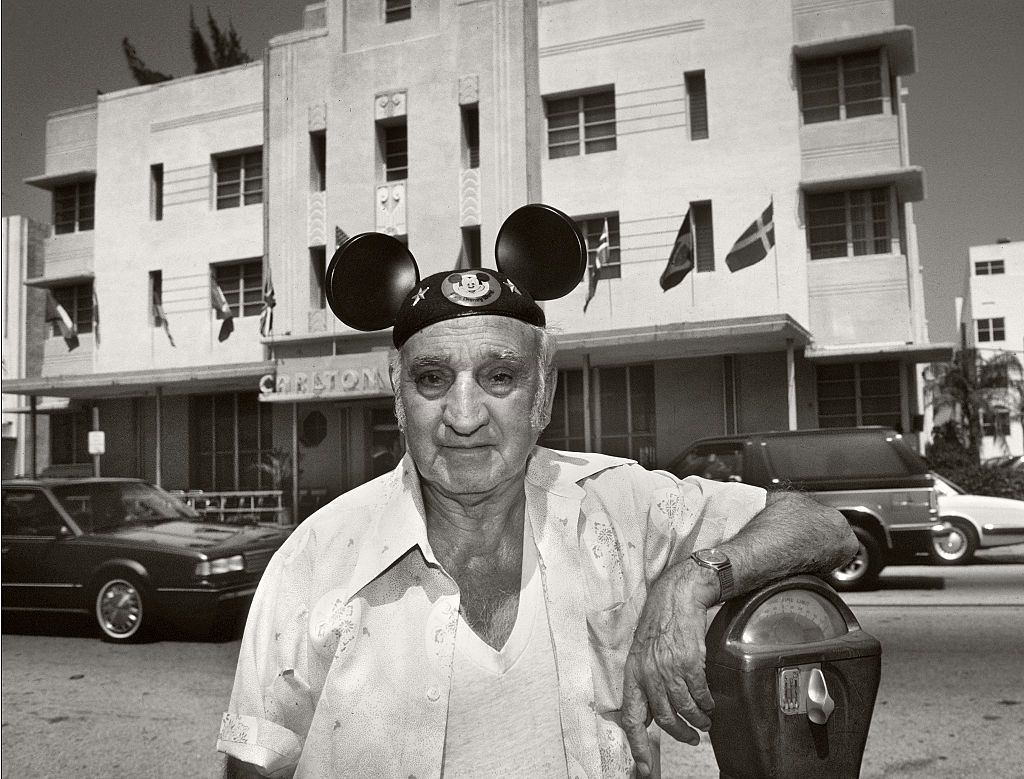 Elderly man standing by a parking meter in front of the Carlton Hotel, Washington Avenue, Miami Beach, wearing a Micky Mouse hat.