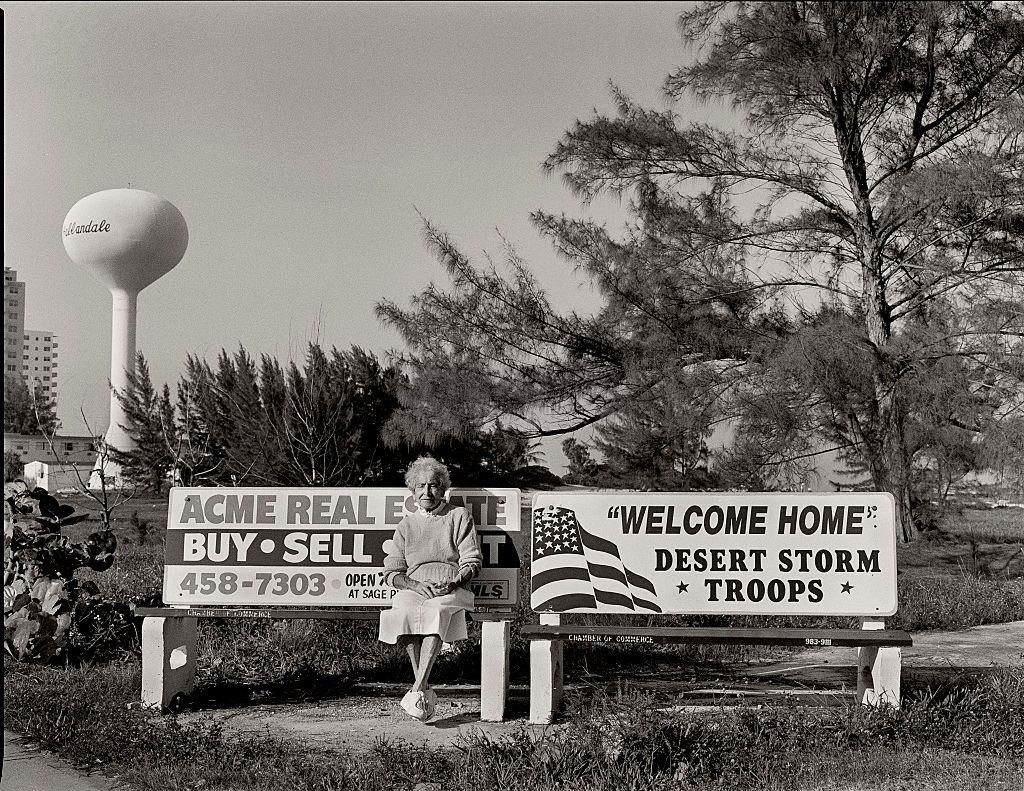 Old woman sitting on street bench with the sign "Welcome home Desert Storm Troops" (from their first venture in Iraq) on Collins Avenue in Sunny Isles, Miami Beach, Florida.