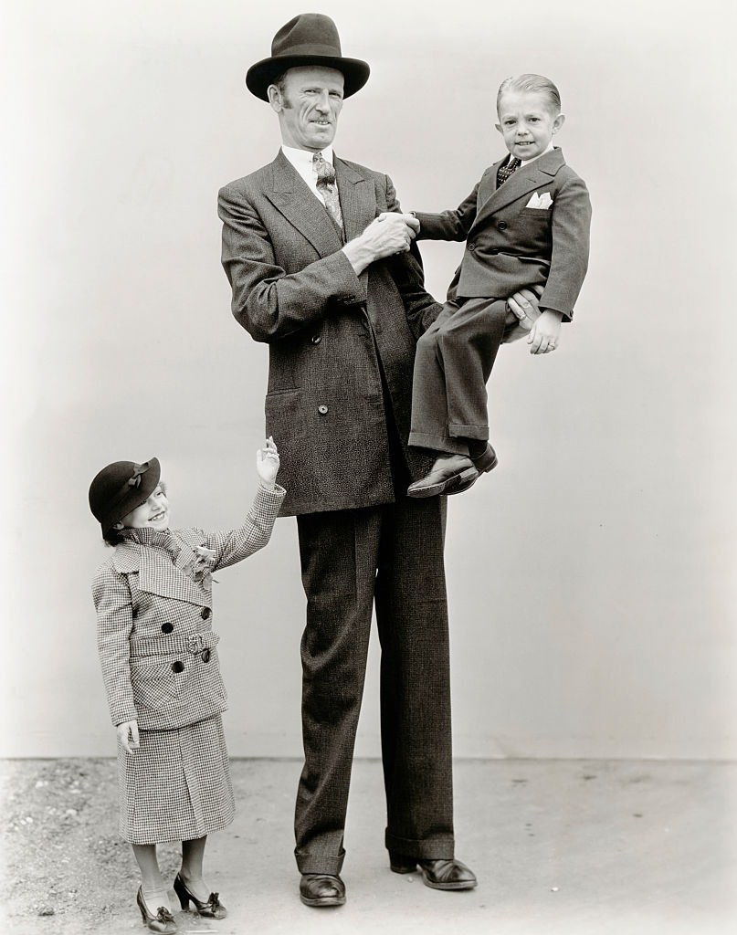 Giant Tex Madsen lifts small person George Brasno off the ground to shake his hand. George's wife Olive waves.