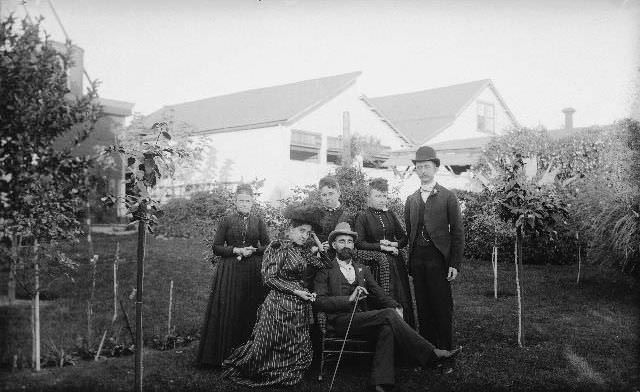 Group of men and women posing for a portrait in the backyard of a house, 1895