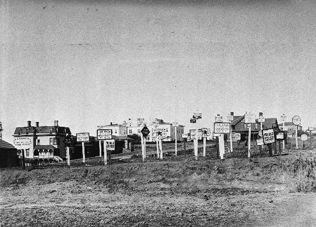 Large number of For Sale signs on a vacant lot with houses in the distance, 1899