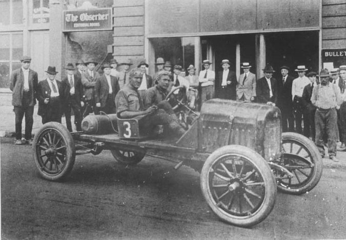 Driver and mechanic pose in their car in front of the Charlotte Observer offices on South Church Street, 1919