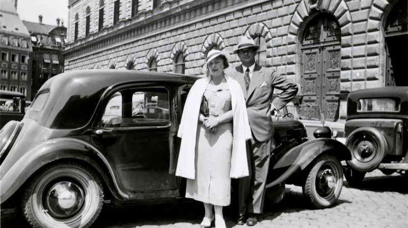An elegant couple posing with a Citroën 11 CV in front of an impressive Renaissance building in a busy historic town centre, circa 1935