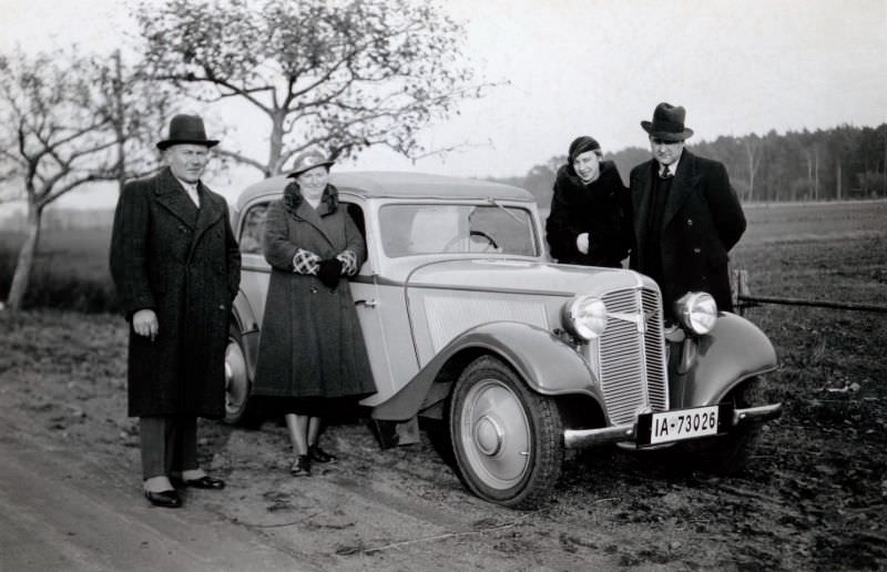 Two stylish couples posing with a Adler Trumpf Junior Cabrio-Limousine on a dirt road in the countryside.