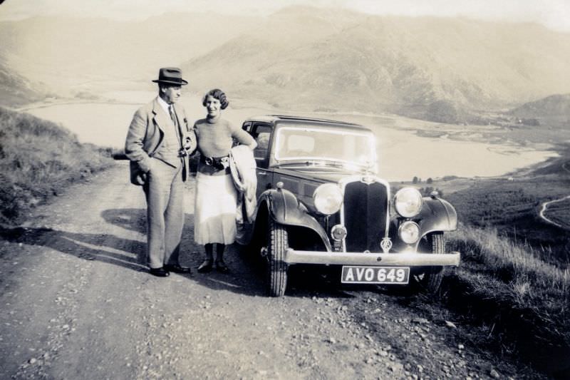 A middle-aged couple posing with a Triumph Gloria Vitesse on a gravel road in the countryside.