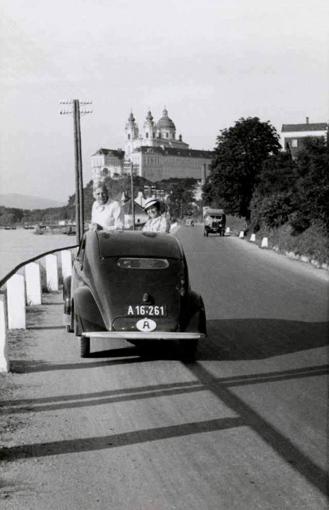 A cheerful couple posing with a Steyr 50 on the side of the road in the town of Melk on the bank of the Danube with the famous monastery visible in the background.