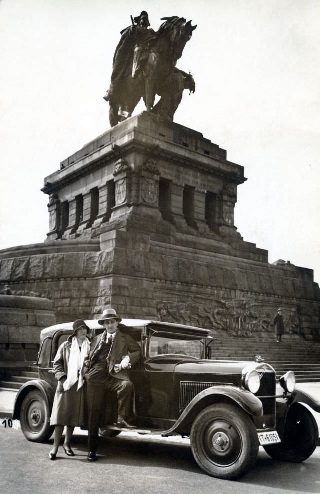 A stylish couple posing with a Röhr 8 9/50 PS Typ R in front of a monumental equestrian statue at Deutsches Eck, 1930