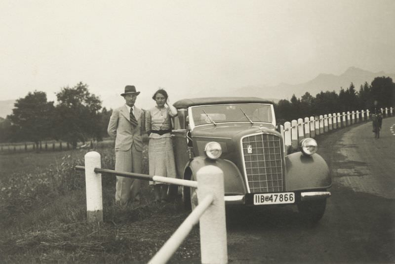 A couple wearing light-colored suits in the fashion of the thirties posing with an Opel 6 Cabriolet in the countryside.