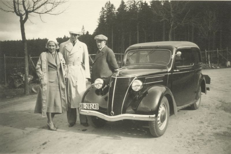 A young elegant couple in trench coats and an elderly gent in a woollen coat posing with a German-built Ford Eifel.