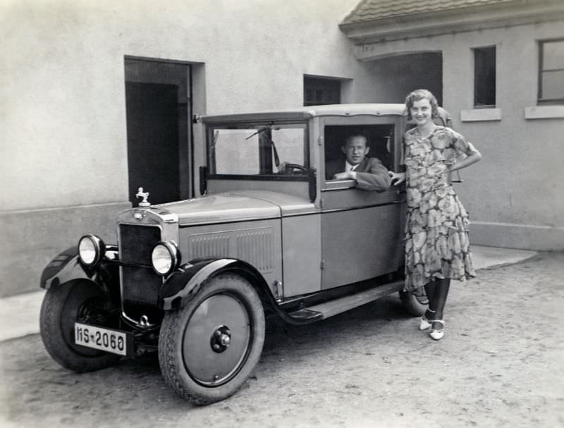 A cheerful lady in a floral dress and a dapper fellow in a suit posing with a Hanomag 4/20 PS Cabriolet-Limousine in the backyard of a private home.