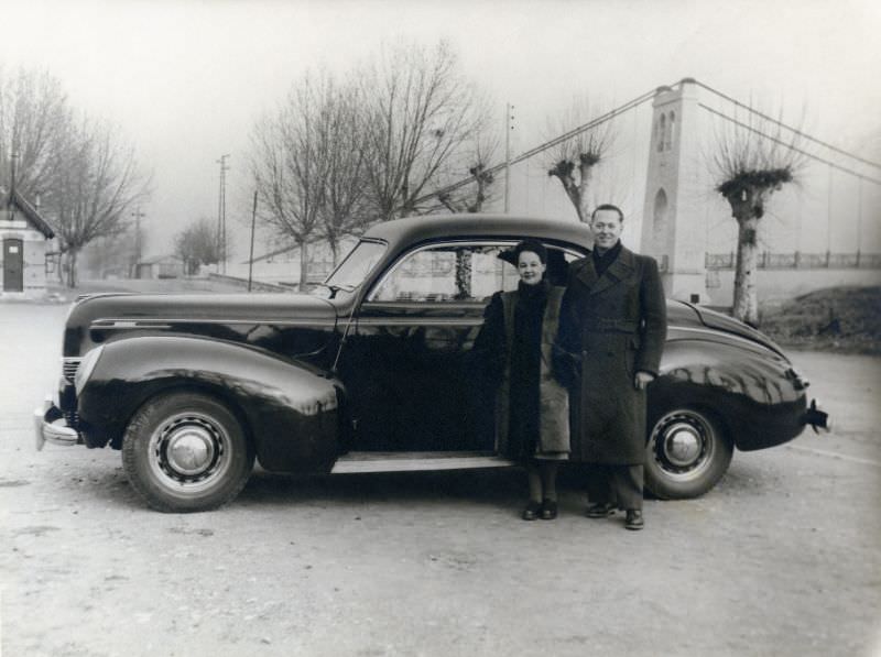 A fashionable couple posing with a 1939 Mercury Eight Club Coupe near a suspension bridge on a bleak winter's day, 1939