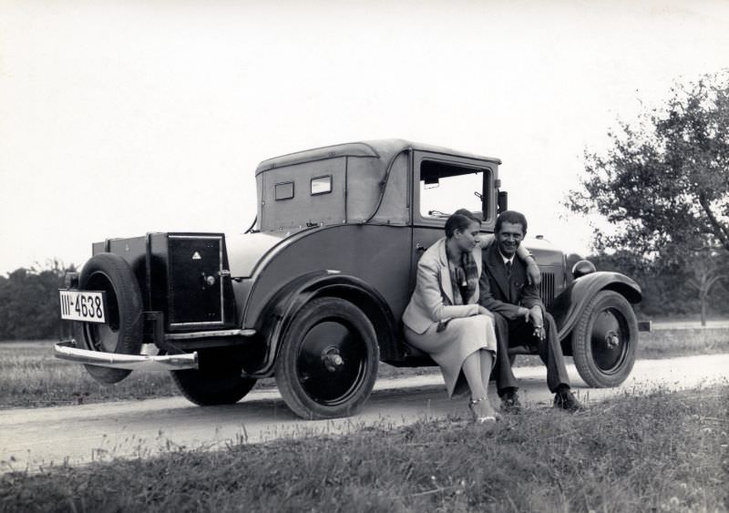 A stylish couple posing on the running board of an Opel 4/20 PS Cabriolet 2 convertible in the countryside.