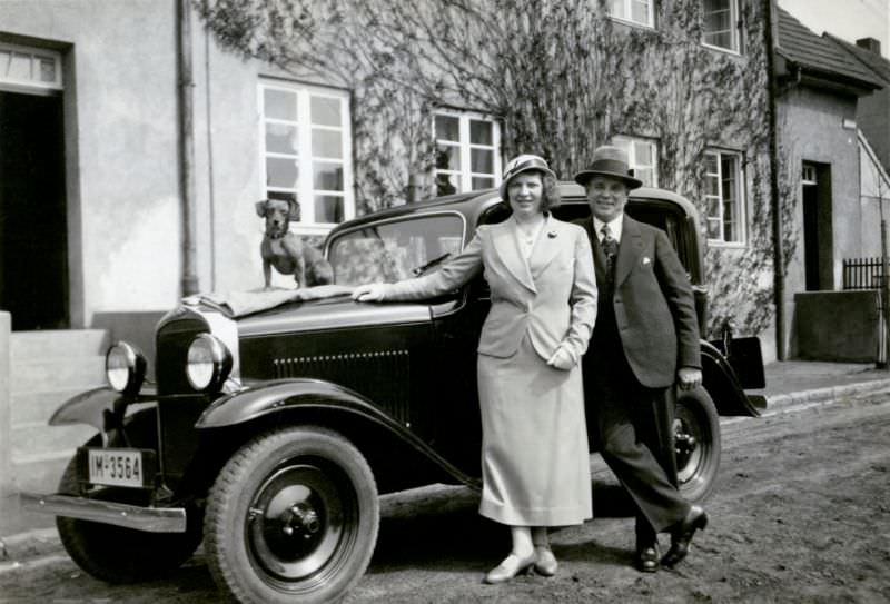 A cheerful couple and posing with an Opel 1,2 Liter in front of a middle-class home in a residential street.