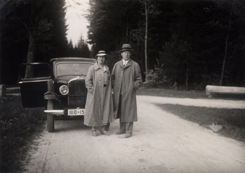 A middle-aged couple wearing raincoats and hats with an Opel 1,2 Liter.