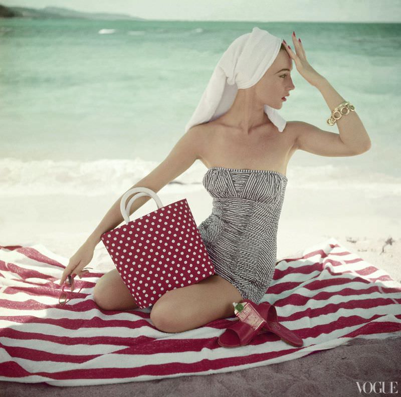 Model Maria Reachi seated on striped beach towel wearing grey and white patterned strapless bathing suit, Vogue, 1954