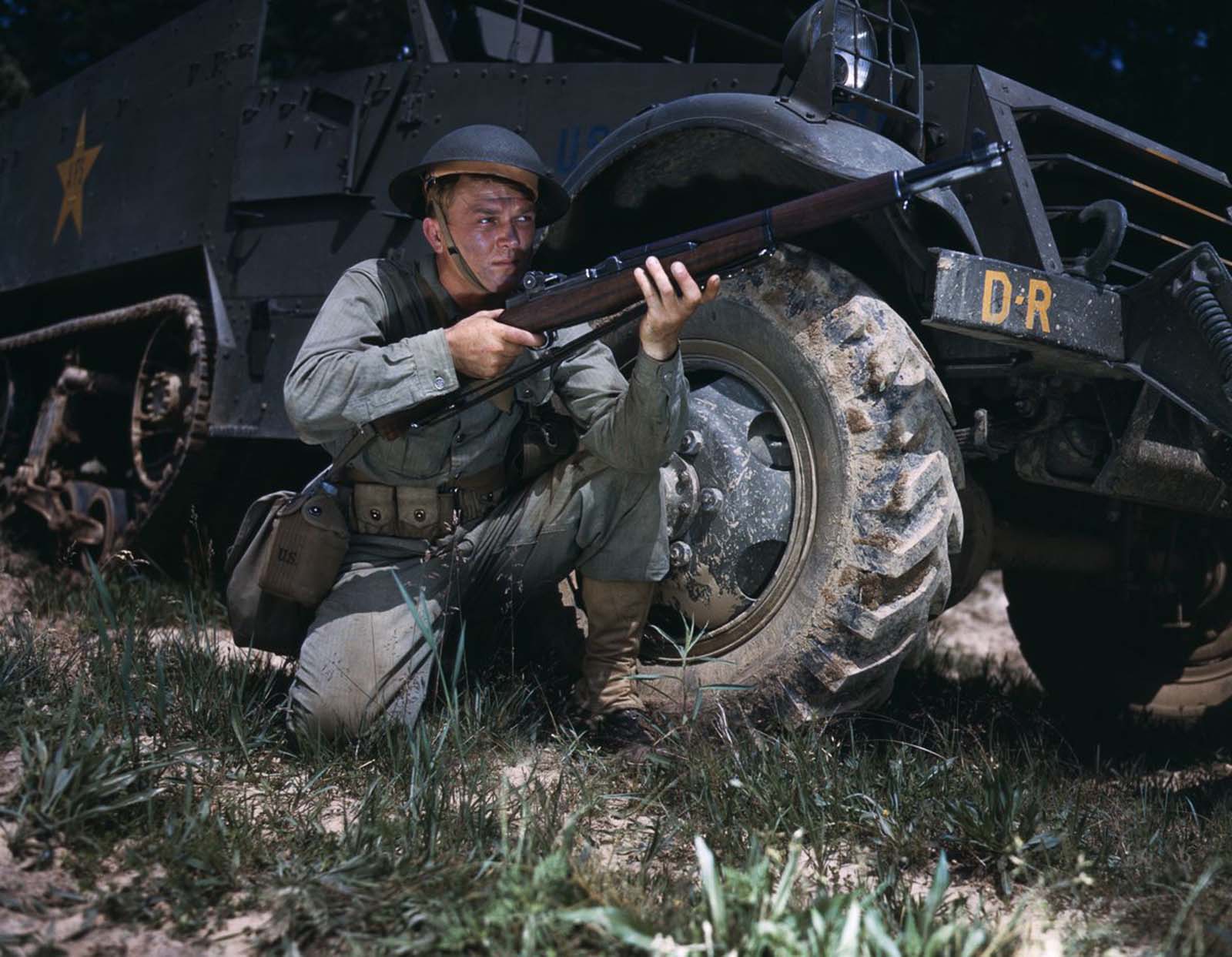 An infantryman crouches beside a half-track with an M1 Garand.