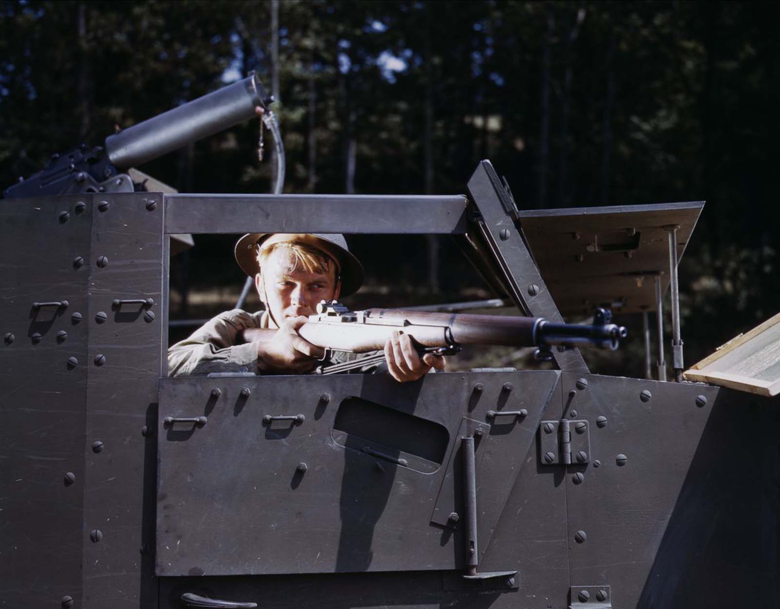 An infantryman aims an M1 Garand from inside a half-track.