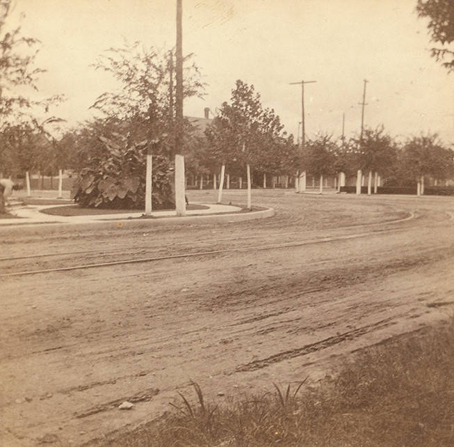 A view of an unidentified dirt street. A man is doing yard work on the left, 1889