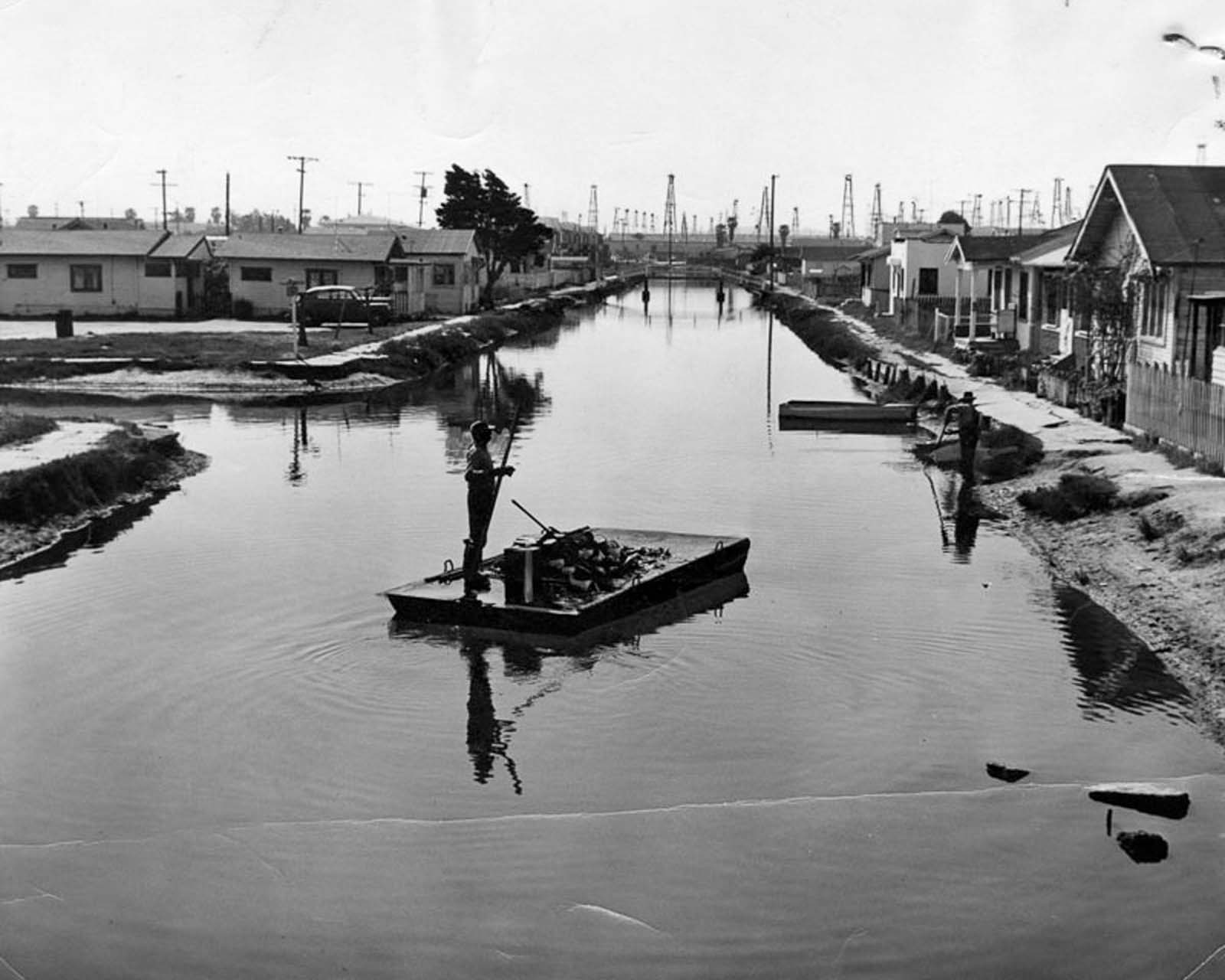 A man poles a garbage scow down the Grand Canal in Venice, California, with oil derricks on the horizon, 1953.
