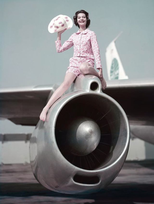 Model sitting atop the engine of Pan Am's 707 Clipper wearing a pink printed sharkskin jacket, 1959