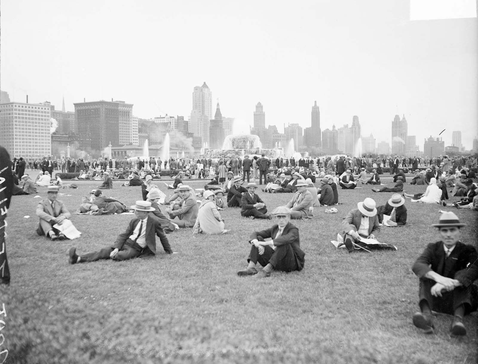 Crowds sitting and standing on a lawn in Chicago’s Grant Park. Buckingham Fountain and buildings are visible in the background.