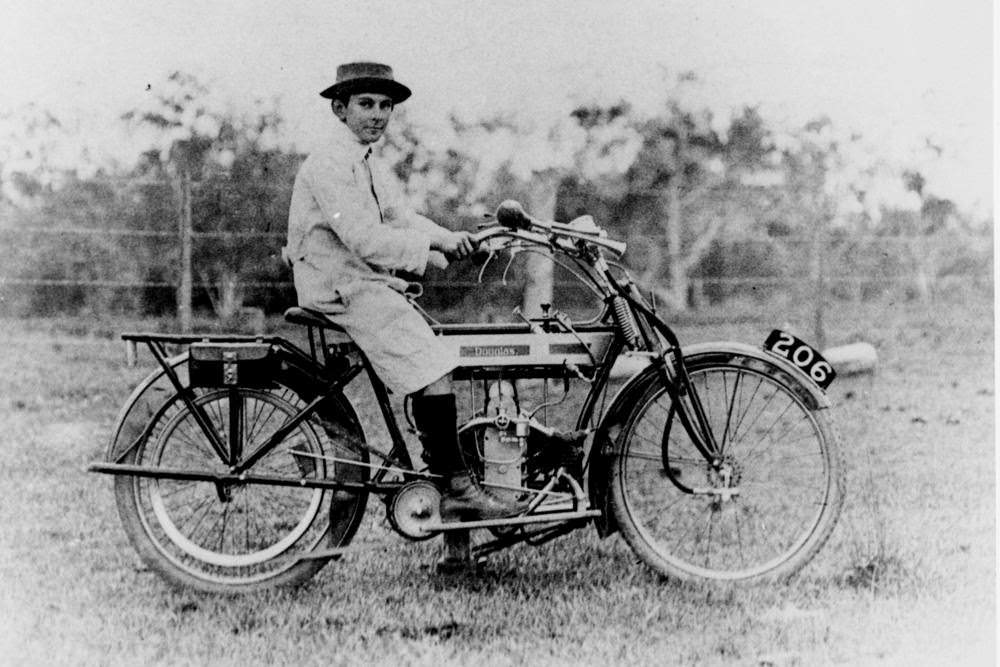A boy, wearing a long coat and a hat with the brim turned up, is sitting on a early model Douglas motorcycle, 1915.