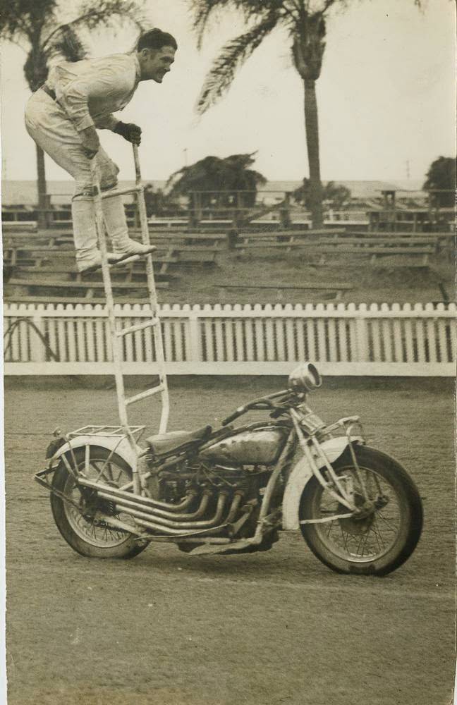 A young man is at the top of a small ladder attached to his motorcycle, 1955.