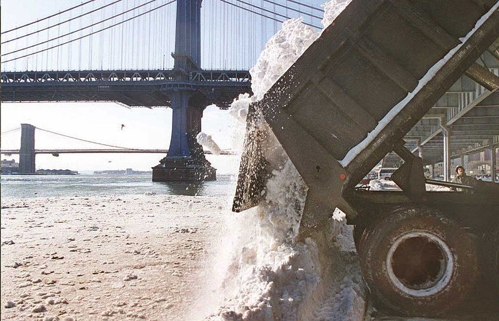 A New York City Department of Sanitation truck dumps snow into the East River underneath the Manhattan Bridge in New York with the Brooklyn Bridge is visible behind.