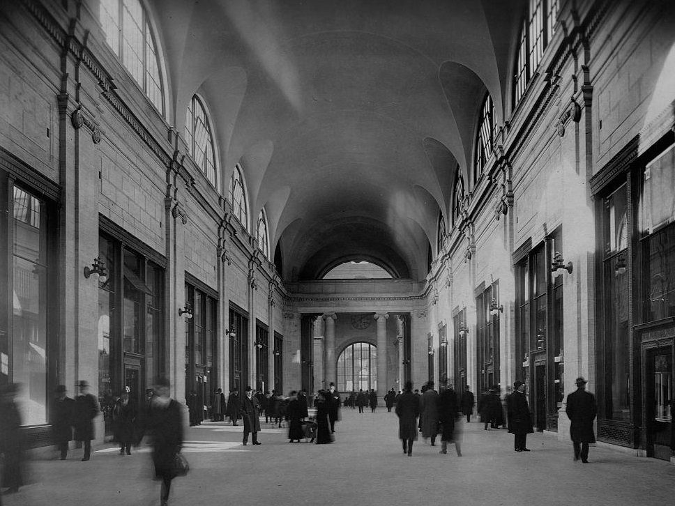 Sunlight cast through the large windows of a vaulted hall in Pennsylvania Station plays across the columns on the wall as commuters below rush to catch their trains., 1911
