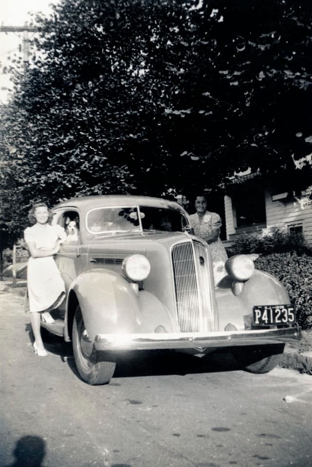Two ladies and their dog posing with a 1936 Studebaker Dictator Six in a suburban street in summertime.