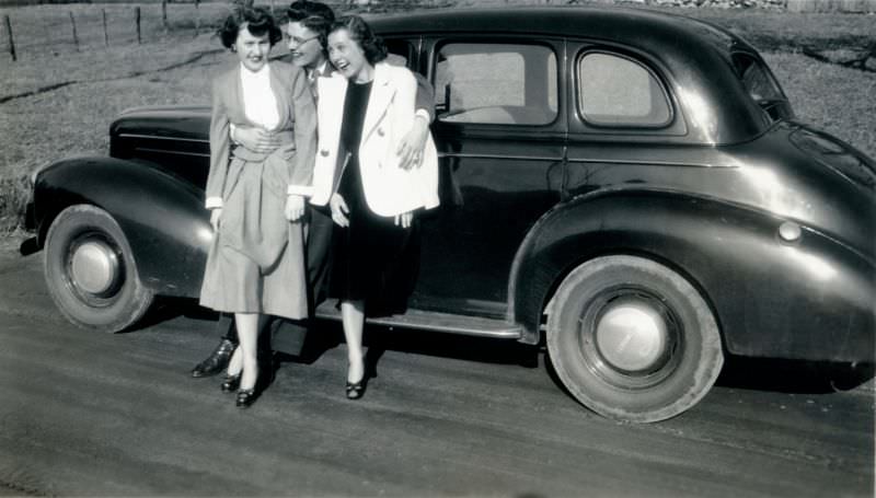 A cheerful company of three – two stylish ladies and a fellow with spectacles – posing with a 1940 Studebaker Sedan on a dirt road, 1948