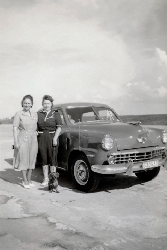 Two middle-aged ladies and their pet Dachshund posing with a 1948 Studebaker Champion in the countryside, 1948