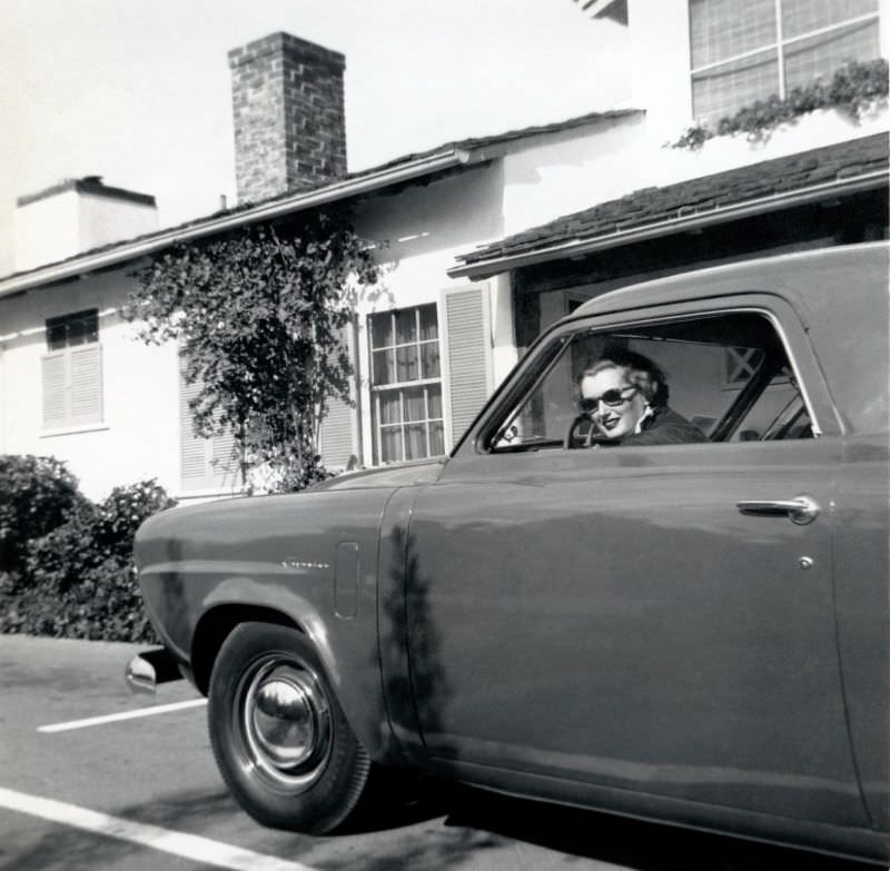 A blonde lady wearing dark sunglasses posing in the driver's seat of a 1950 Studebaker Champion in front of a suburban home, 1950