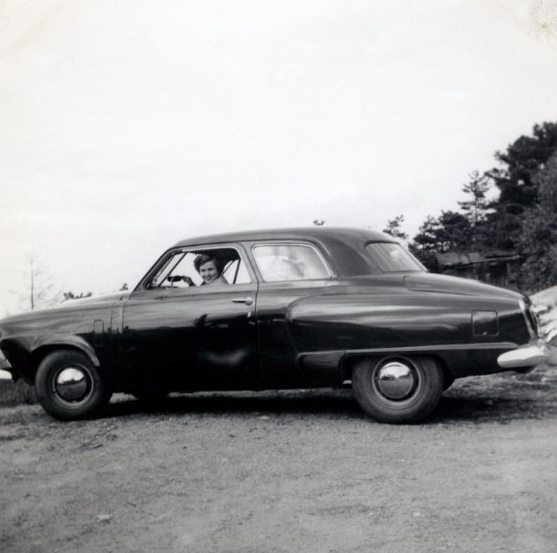 A cheerful lady posing in the driver's seat of a 1950 Studebaker Champion Custom 2-Door Sedan in a gravel parking lot on the outskirts of town, 1950