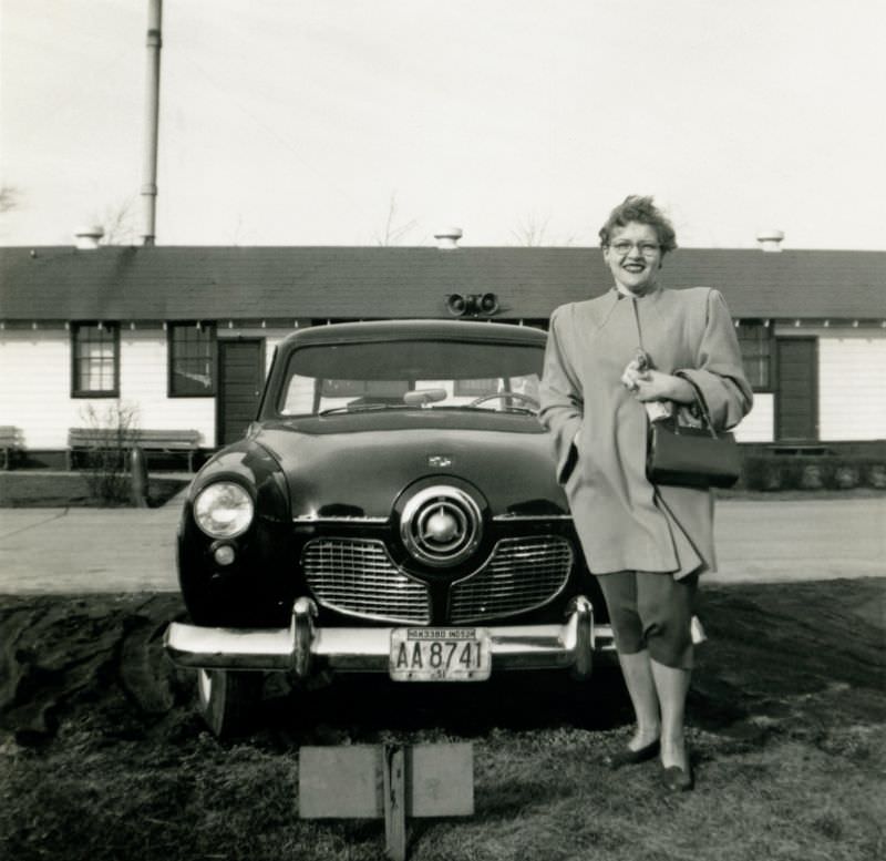 A middle-aged lady posing with a 1951 Studebaker Champion Custom in front of a timber house.