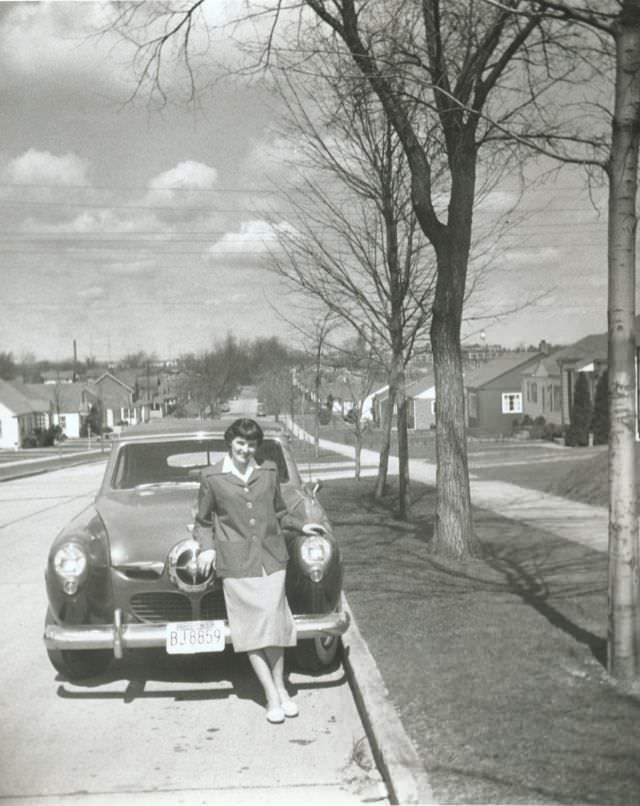 A young woman posing with a 1950 Studebaker Champion Convertible in a suburban street on a sunny winter's day, 1952