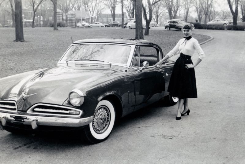 A cheerful young lady posing with a 1953 Studebaker Commander Starliner in a suburban street in autumn time, 1953