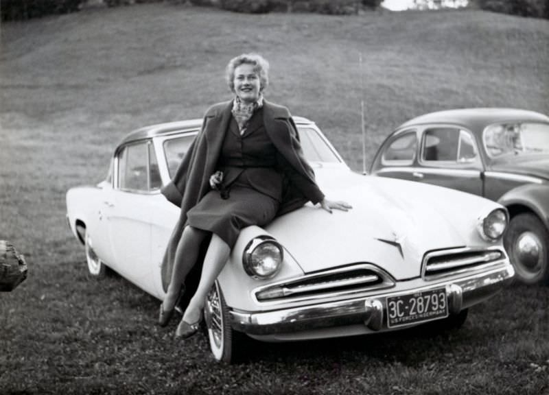 A platinum-blonde lady posing on the fender of a 1953 Studebaker Champion Regal Starliner Coupe.