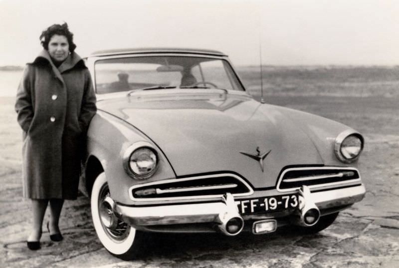A middle-aged lady in a woollen coat posing with a 1953 Studebaker Champion Regal Starliner in wintertime.