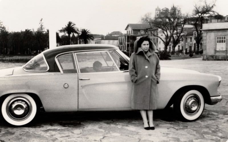 A slightly grumpy-looking lady in a woolen coat posing with a 1953 Studebaker Champion Regal Starliner on a bleak winter's day.