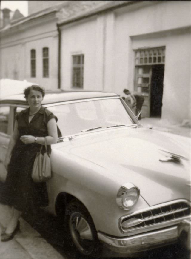 A lady in a short sleeved dress posing with a 1954 Studebaker Sedan in a residential street, 1950s