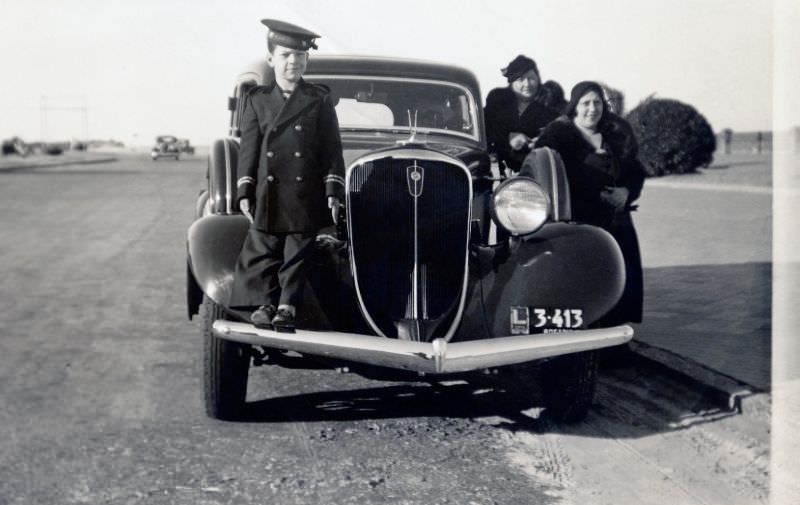 A little boy in a sailor's uniform standing on the bumper of a 1934 Studebaker Land Cruiser.