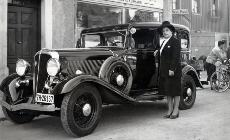 A middle-aged lady in a dark female suit posing with a 1933 Rockne (Studebaker) “10” Deluxe 4-door Sedan in a residential street.