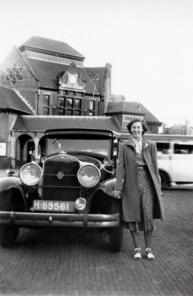 A lady wearing a floral dress, woollen coat, and white sandals posing with a 1929-30 Studebaker Eight Sedan in a busy cobbled street, 1938