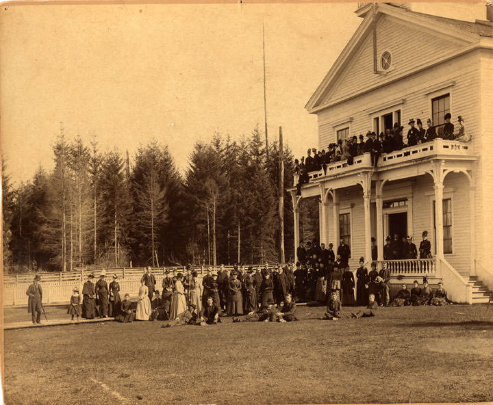 A large group of men and women, and at least one child, in front of and on the balcony and porch of Olympia, 1889.