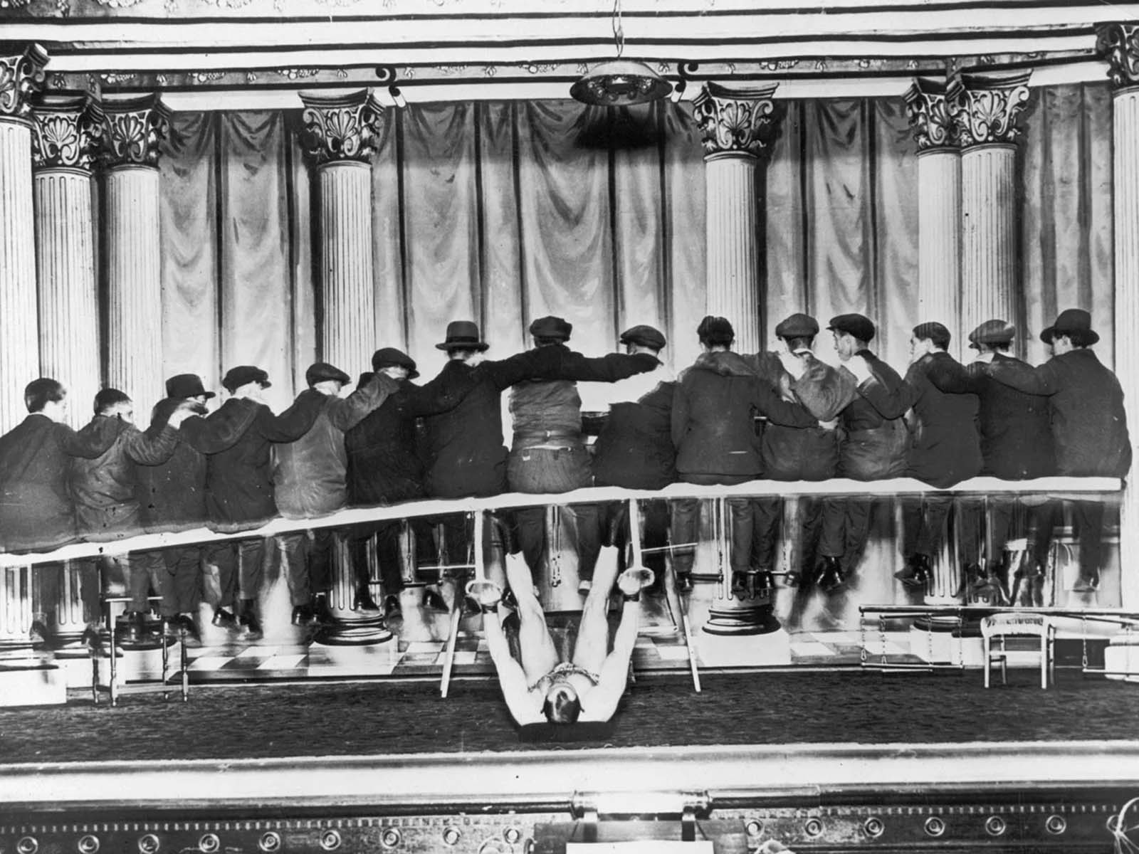 A strongman lying on his back onstage, lifting a group of men and the bench on which they sit, 1935