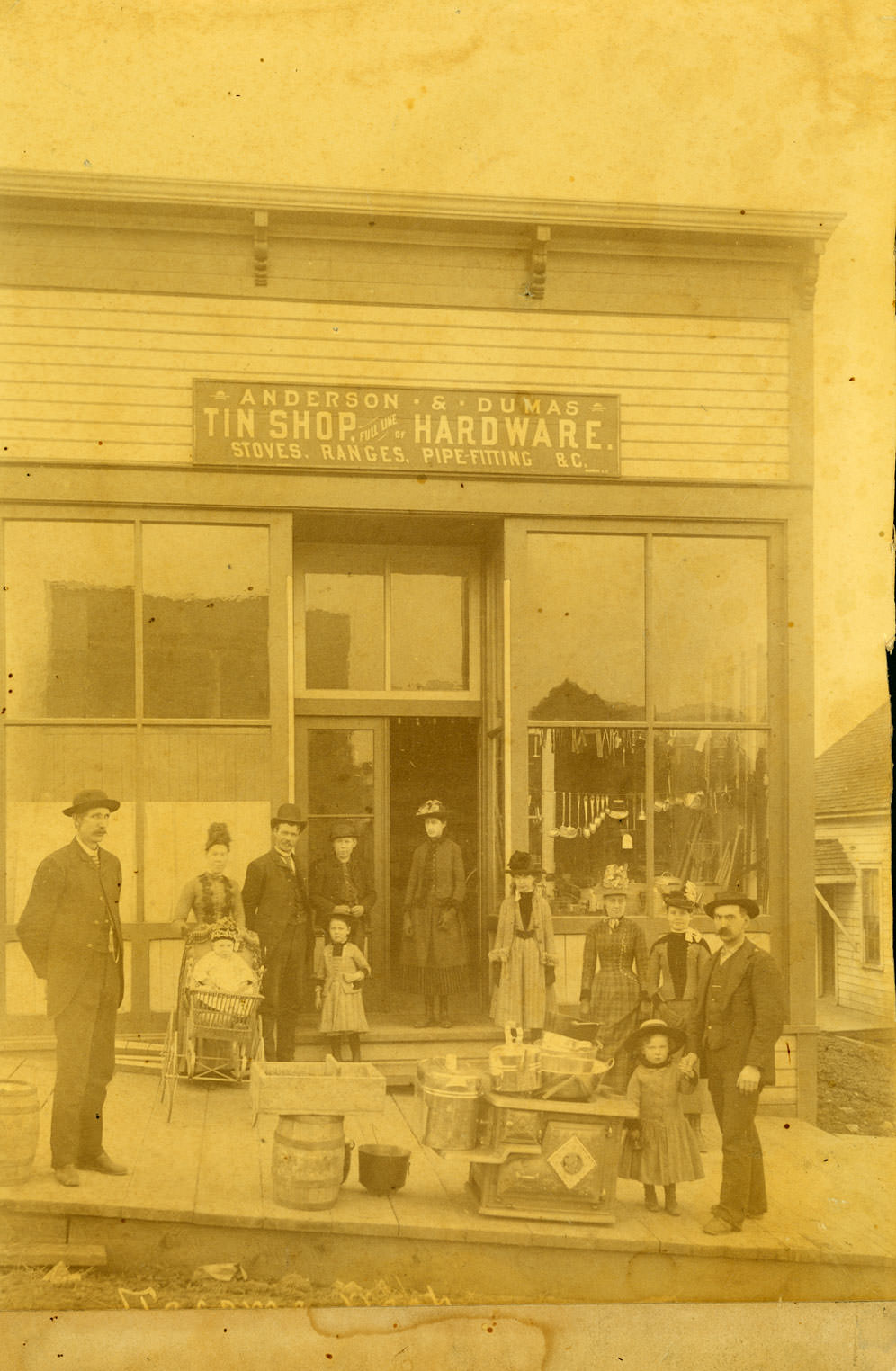 Six women, three men, and three girls, one in a carriage, stand in front of the Anderson and Dumas Tin Shop at Second and McCarver Streets, Tacoma, 1888