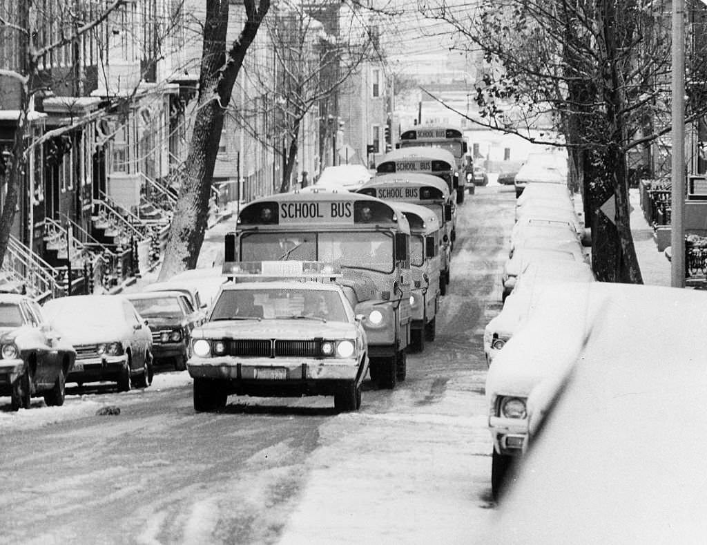On a snowy day, school buses follow a police car along G Street to South Boston High School in Boston, 1974.